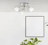 A ceiling light fixture with three adjustable spotlights is mounted on a white ceiling above a framed piece of art and a plant. Produktbild-Vorschau