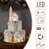 A decorative illuminated Christmas village scene with a tower and multiple houses, displayed on a wooden surface, with a Christmas tree and ornaments in the background. Produktbild-Vorschau