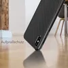 A close-up, angled shot of a black carbon fiber-look phone case on a wooden floor, with a partial view of a wooden post and a chair in the background. Produktbild-Vorschau