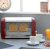 A red toaster with bread inside, next to two cups of coffee and a glass of orange juice on a kitchen counter. Podgląd zdjęcia produktu