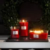 Three red WoodWick candles of varying sizes are displayed on a dark slate surface in front of a green wreath. Podgląd zdjęcia produktu