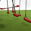 A close-up view of a red metal swing set with green artificial turf flooring. Produktbild-Vorschau