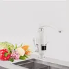 A white electric hot water tap is shown in a kitchen setting next to a selection of fresh vegetables. Náhled obrázku produktu