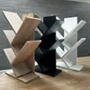 Three tree-shaped bookshelves in different colors (wood tone, black, and white) are displayed on a wooden table against a gray tiled wall. Náhľad obrázku produktu