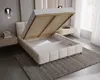 A modern bedroom with a cream bouclé upholstered bed frame, its storage compartment open, revealing a slatted mattress base and storage space. A bedside table with books and a lamp is visible to the left, and sheer curtains frame a window to the right. Náhled obrázku produktu