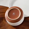 The underside of a ceramic bowl with a logo and text, resting on a wooden surface next to a white plate. Produktbild-Vorschau