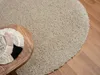 A round, beige shaggy rug is placed on a wooden floor next to a wooden table with a decorative placemat. Náhľad obrázku produktu