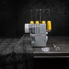 A sewing machine with yellow thread spools sits on a dark metallic surface, with a piece of dark fabric draped over the left side. Produktbild-Vorschau