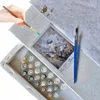 A hand holding a paintbrush is about to paint on a canvas, with paints and a reference image laid out. Produktbild-Vorschau