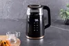 A black electric kettle with a digital display sits on a countertop next to a glass cup and a pastry. Náhled obrázku produktu