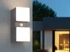 An outdoor wall light with a motion sensor is mounted on a gray wall next to a modern house. Anteprima dell’immagine prodotto