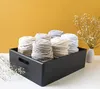 A black wooden crate filled with spools of thick yarn in various neutral colors, placed on a white surface against a yellow wall. A small vase with dried flowers is visible to the right. Produktbild-Vorschau