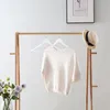 A cream-colored sweater on a wooden hanger, hanging on a wooden rack. There is a straw hat on the rack, and a vase of flowers in the foreground. Produktbild-Vorschau