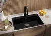 A modern kitchen sink with a black granite basin and a black faucet, set against a slatted wooden backsplash. A pitcher of green liquid and glasses are on a tray to the left, and a bowl of fruit is on the counter to the right. Produktbild-Vorschau