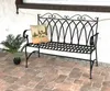 A black metal garden bench with a decorative backrest, placed on a brick patio next to a potted plant and a decorative pillow. Anteprima dell’immagine prodotto