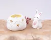 A white ceramic candle holder with star cutouts and a small white bunny figurine with pink accents are displayed on a wooden surface. Anteprima dell’immagine prodotto