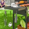 A person is sitting on the grass in the background while a gas grill is in the foreground with various cooking utensils and vegetables. Náhled obrázku produktu