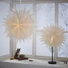 Two white paper star lanterns are displayed on a windowsill in front of a window showing a snowy outdoor scene. One is hanging, and the other is on a black stand. Cinnamon sticks and twine are on the windowsill. Podgląd zdjęcia produktu
