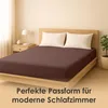 A bed with a brown fitted sheet and two white pillows, set in a bedroom with a nightstand and a plant. Produktbild-Vorschau
