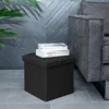 A dark gray fabric storage ottoman sits on a tiled floor next to a dark gray sofa. Two books are stacked on top of the ottoman. A large green leaf plant is in the upper left corner. Náhľad obrázku produktu