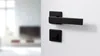 A black door handle and lock mechanism are shown mounted on a white wall, with a blurred interior scene in the background. Produktbild-Vorschau