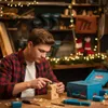 A man is carving a wooden star in a workshop decorated with Christmas lights. Anteprima dell’immagine prodotto