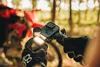 A person's hands wearing black gloves are holding a GoPro camera mounted on a bicycle handlebar. A red bicycle is visible in the blurred background. Anteprima dell’immagine prodotto