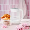 A glass kettle is shown on a table with croissants and a cup of tea, decorated with pink flowers, in front of a pink tiled wall. Produktbild-Vorschau