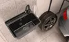 A black sink is shown next to a car tire and a car in a garage or workshop setting. Produktbild-Vorschau
