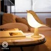 A bird-shaped lamp sits on a marble table next to a book, with a blurred background of a living room. Anteprima dell’immagine prodotto