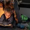 A young boy is reading a book in a tent illuminated by string lights, with a dragon-shaped night light on a stack of books nearby. Produktbild-Vorschau