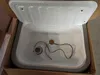 A white sink with a gray rim is shown inside a cardboard box. The lid of the sink is open. Náhled obrázku produktu