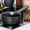 A black saucepan with a speckled interior sits on a reflective black surface, with jars of coffee beans and granola in the background. Náhled obrázku produktu