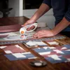 A person is using a rotary cutter to cut fabric on a wooden table, with pre-cut fabric pieces laid out. Produktbild-Vorschau