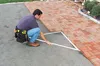 A man is kneeling on a gravel surface next to a brick pathway, using a triangular measuring tool. Náhled obrázku produktu