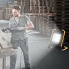 A man is working in a warehouse with stacks of wooden pallets, using a reciprocating saw on a piece of wood. A portable LED work light is illuminating the scene. Náhled obrázku produktu