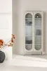 A beige display cabinet with glass doors and arched tops, placed in a minimalist room with a vase of dried flowers and a piece of art on the wall. Voir les images du produit