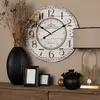 A decorative wall clock is displayed on a wooden shelf, surrounded by other decorative items like vases and picture frames. Produktbild-Vorschau