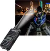 A person in a high-visibility vest and beanie crouches down at night, illuminated by a bright light, with a walkie-talkie in the foreground. Produktbild-Vorschau