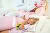 A young girl is sleeping on a bed, hugging a large pink plush unicorn. Other plush unicorns are visible in the background. Podgląd zdjęcia produktu