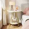 A round side table with a lamp, books, and a small basket next to a bed. Produktbild-Vorschau