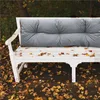 A white garden bench with a gray cushion, covered in fallen autumn leaves. Náhled obrázku produktu