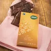 A close-up shot of a chocolate bar box and several broken chocolate pieces on a pink cloth and a wooden surface. Voir les images du produit