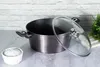 A dark gray pot with a glass lid is placed on a light gray countertop, with a small white bowl of white powder to the left and a green plant to the right. The background is a white brick wall. Produktbild-Vorschau