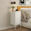 A white bedside table with a closed door and a small vent detail on the lower front is placed next to a bed. On top of the table are a few books, a camera, and a small vase with greenery. A wall lamp is mounted on the wall above the table. Anteprima dell’immagine prodotto