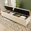 A beige upholstered storage bench with its lid open, revealing stored items like blankets and pillows. The bench is placed on a rug in a room with a window and a plant. Anteprima dell’immagine prodotto