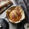 A loaf of bread is placed in a Dutch oven, with a rolling pin and flour on a cutting board in the background. Produktbild-Vorschau