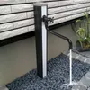 A black outdoor faucet with a curved spout is installed next to a white brick wall, with water flowing into a bed of dark gray pebbles. Podgląd zdjęcia produktu
