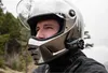 A man wearing a motorcycle helmet with a communication system attached, outdoors with a natural background. Podgląd zdjęcia produktu