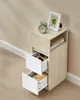 A narrow wooden bedside table with two white drawers and an open shelf is placed against a beige wall and wooden floor. Náhled obrázku produktu
